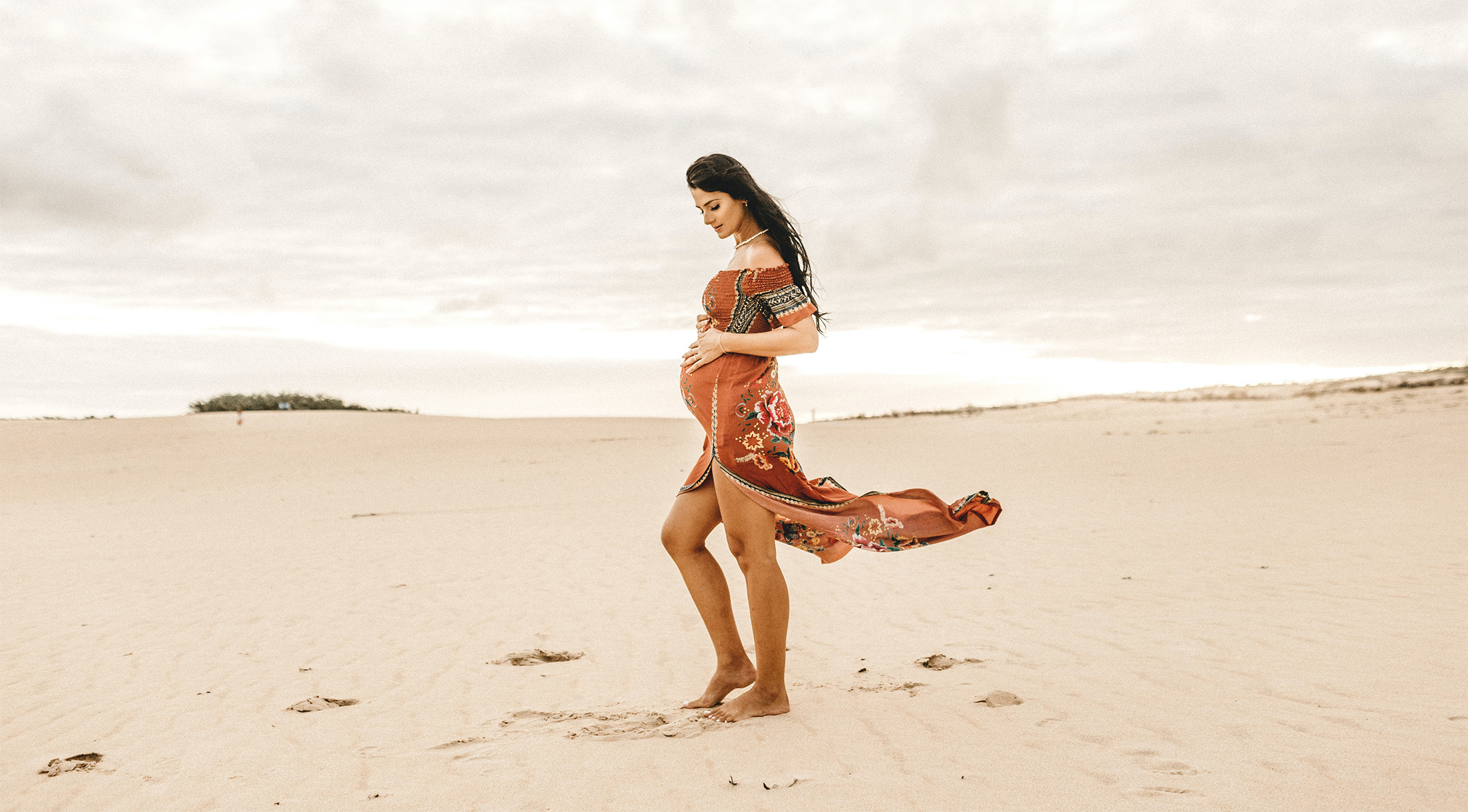 Kobieta w ciąży stoi na plaży i trzyma się za brzuch. Wiatr rozwiewa jej suknię