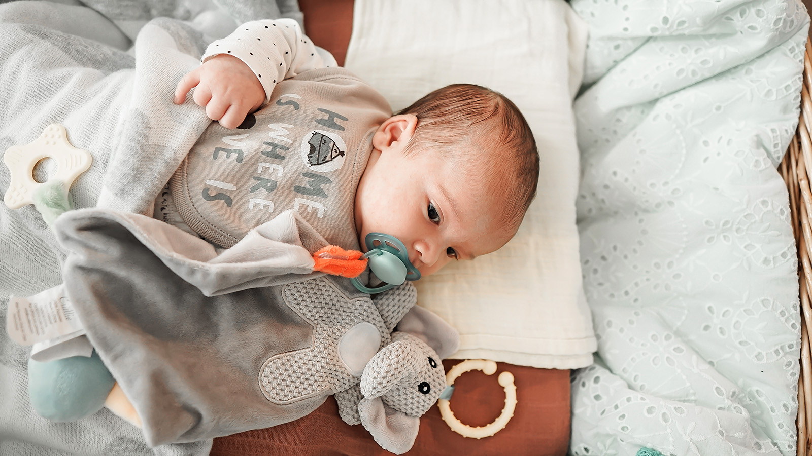 Niespokojny sen niemowlaka może mieć różne przyczyny, zarówno fizjologiczne, jak i środowiskowe - na zdjęciu niemowlę leży na łóżku, pod głową ma białą pieluszkę, w buzi trzyma smoczek, obok leży maskotka HelloMama
