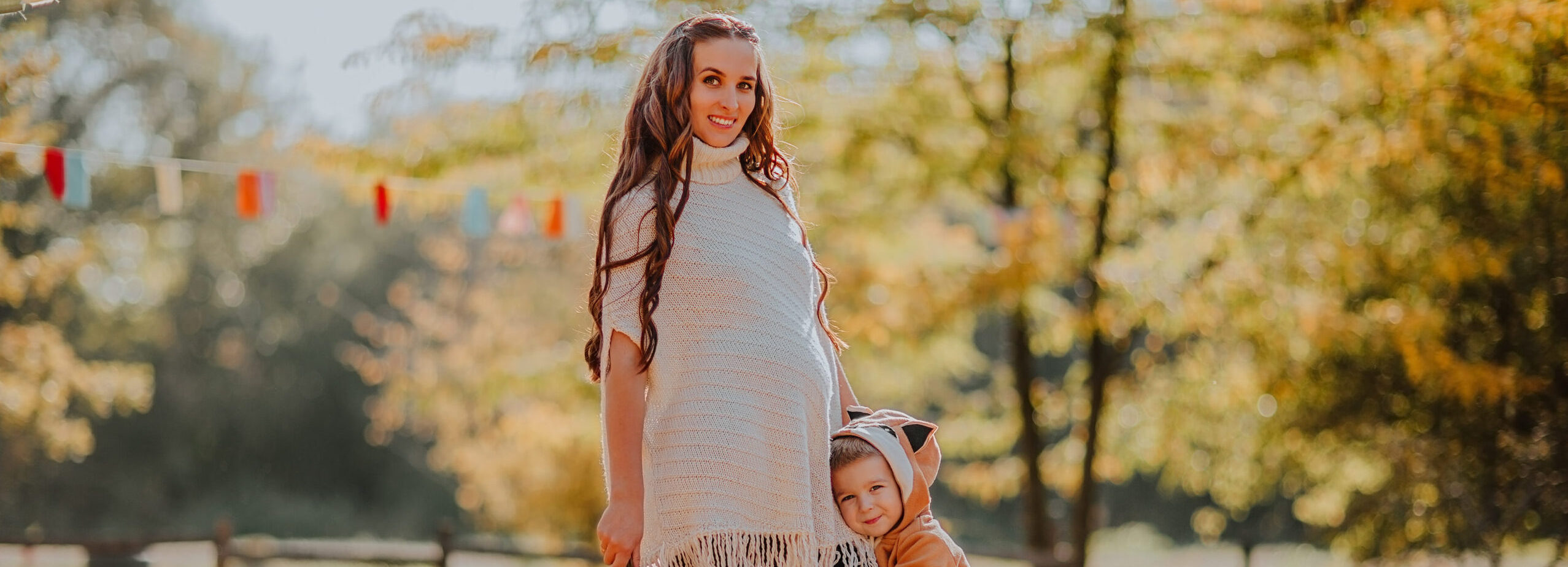 Young pregnant woman in white autumn cardigan and red boots posing with a little son in fox coat at the pumpkin's field. Fall background.